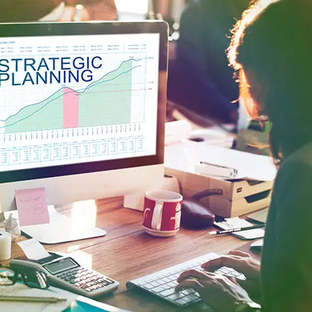 Eine Person sitzt an einem Schreibtisch und tippt auf einer Tastatur, während sie an einer WordPress-Website arbeitet. Auf einem Computerbildschirm wird ein Diagramm mit dem Titel „Strategische Planung“ angezeigt. Der Schreibtisch ist mit Papieren, einer rot-weißen Tasse, Haftnotizen und Büromaterial übersät. Helles Sonnenlicht fällt in den Raum.