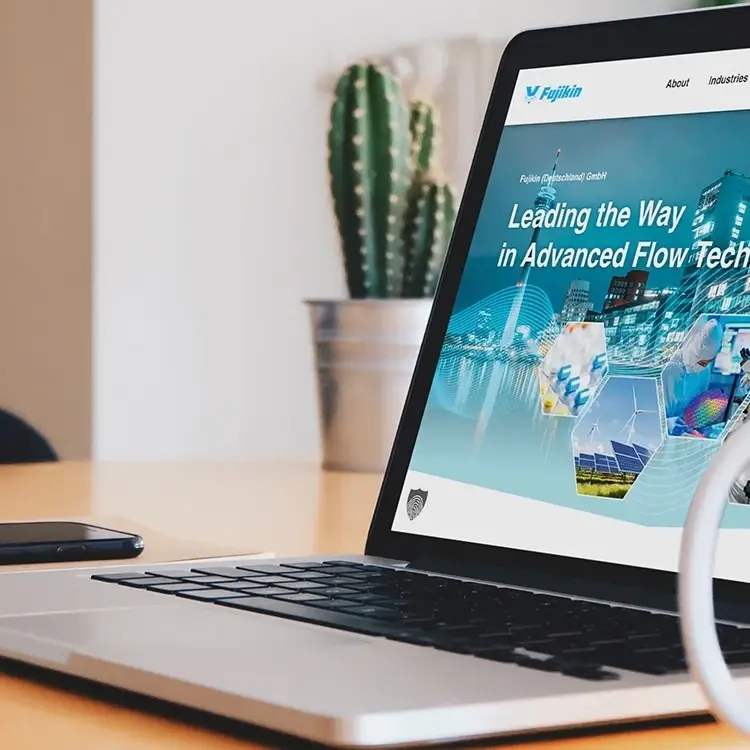 Auf einem Holzschreibtisch steht ein Laptop mit der Firmenhomepage und dem Text „Leading the Way in Advanced Flow Technology“. Daneben sorgt ein Kaktustopf für Grün, während der Teil einer kabellosen Maus zu sehen ist – ein Beweis für durchdachtes Webdesign in Düsseldorf.