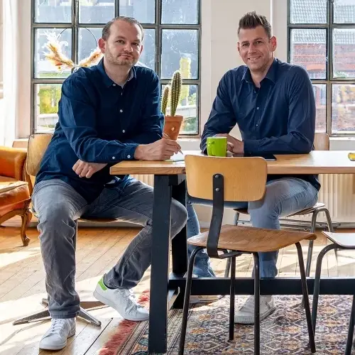 Zwei Männer sitzen an einem Holztisch in einem gut beleuchteten Raum mit großen Fenstern und diskutieren möglicherweise die neuesten Trends im Webdesign in Düsseldorf. Beide tragen dunkle Hemden, auf dem Tisch stehen ein kleiner Kaktus im Topf und eine grüne Tasse. Auf dem Holzboden ist teilweise ein Teppich zu sehen.