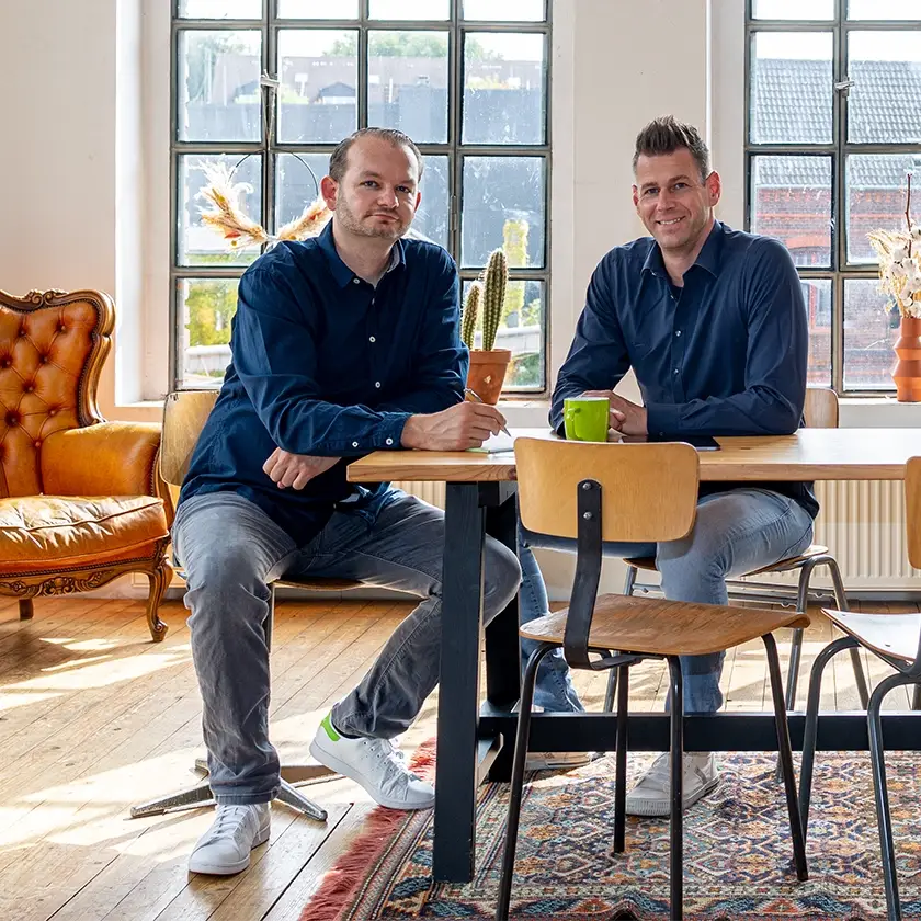 Zwei Männer in legerer Kleidung diskutieren an einem Holztisch in einem hellen Raum mit großen Fenstern über eine WordPress-Website. Der Mann links ruht mit dem Arm auf dem Tisch, umgeben von Stühlen, einem Kaktus und einem Ledersessel. Ein bunter Teppich verstärkt die gemütliche Atmosphäre.