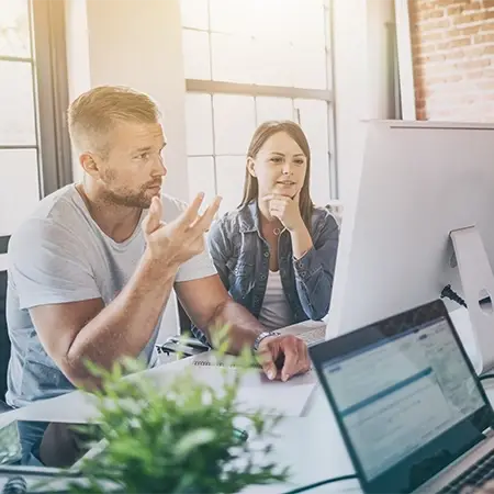 Zwei Personen sitzen an einem Schreibtisch und arbeiten konzentriert an Webdesign in Düsseldorf. Der Mann gestikuliert beim Sprechen auf den Bildschirm, die Frau sitzt nachdenklich da und hat die Hand am Kinn. Im Vordergrund ist ein geöffneter Laptop mit einer Firmenhomepage zu sehen, daneben schmückt eine Pflanze den Arbeitsplatz.