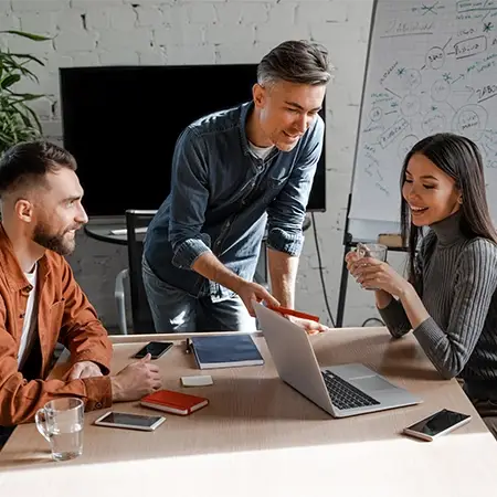 Drei Personen diskutieren angeregt in einem Besprechungsraum über etwas. Eine Person gestikuliert im Stehen und hebt Ideen zum Thema Webdesign Düsseldorf hervor, während zwei andere mit einem Laptop sitzen. Im Hintergrund ist ein Whiteboard mit Schrift zu sehen, umgeben von verschiedenen Notizbüchern und Geräten auf dem Tisch.